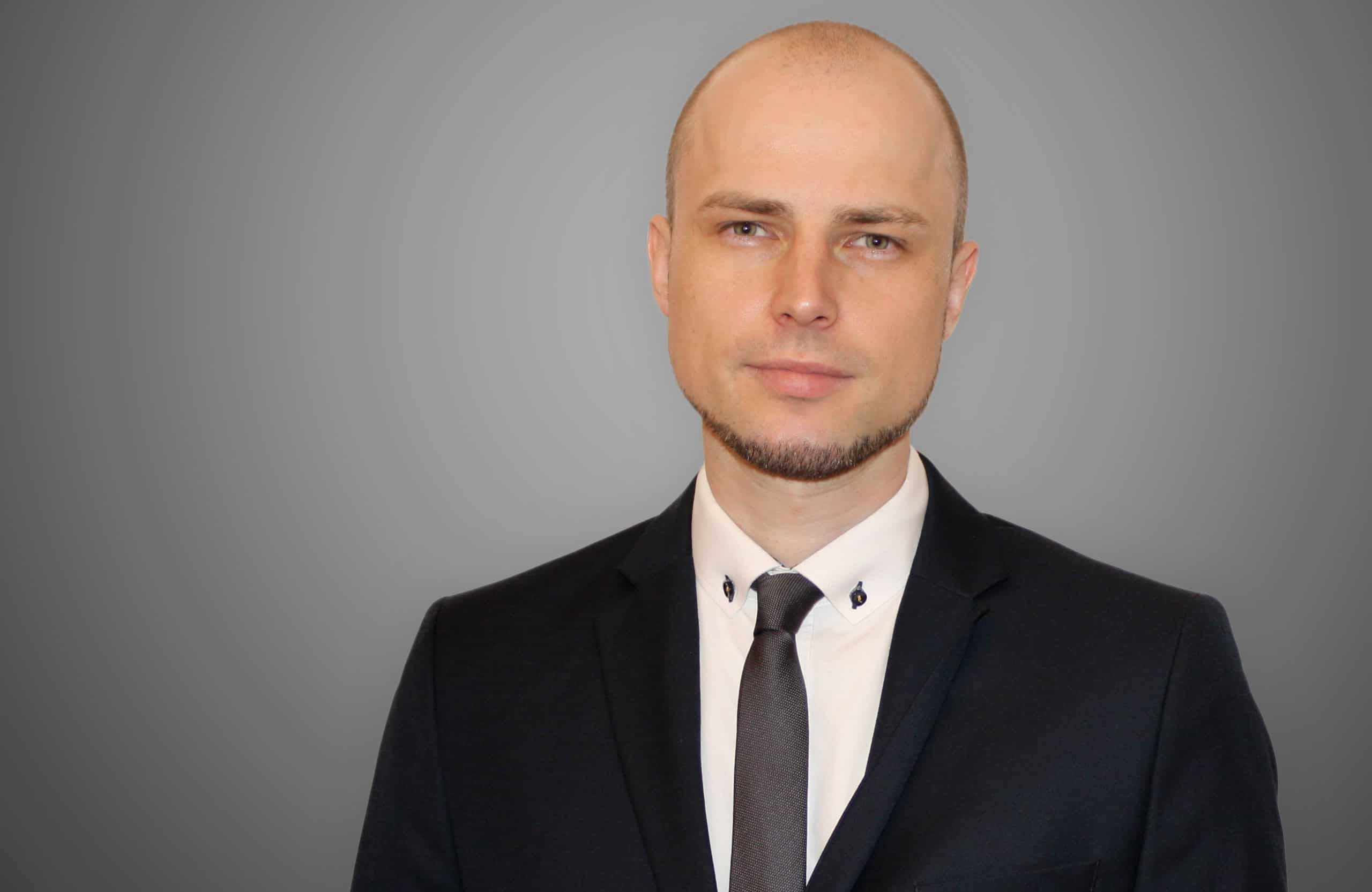 Der Übersetzer Martin Rother trägt einen Anzug und steht vor einer grauen Wand