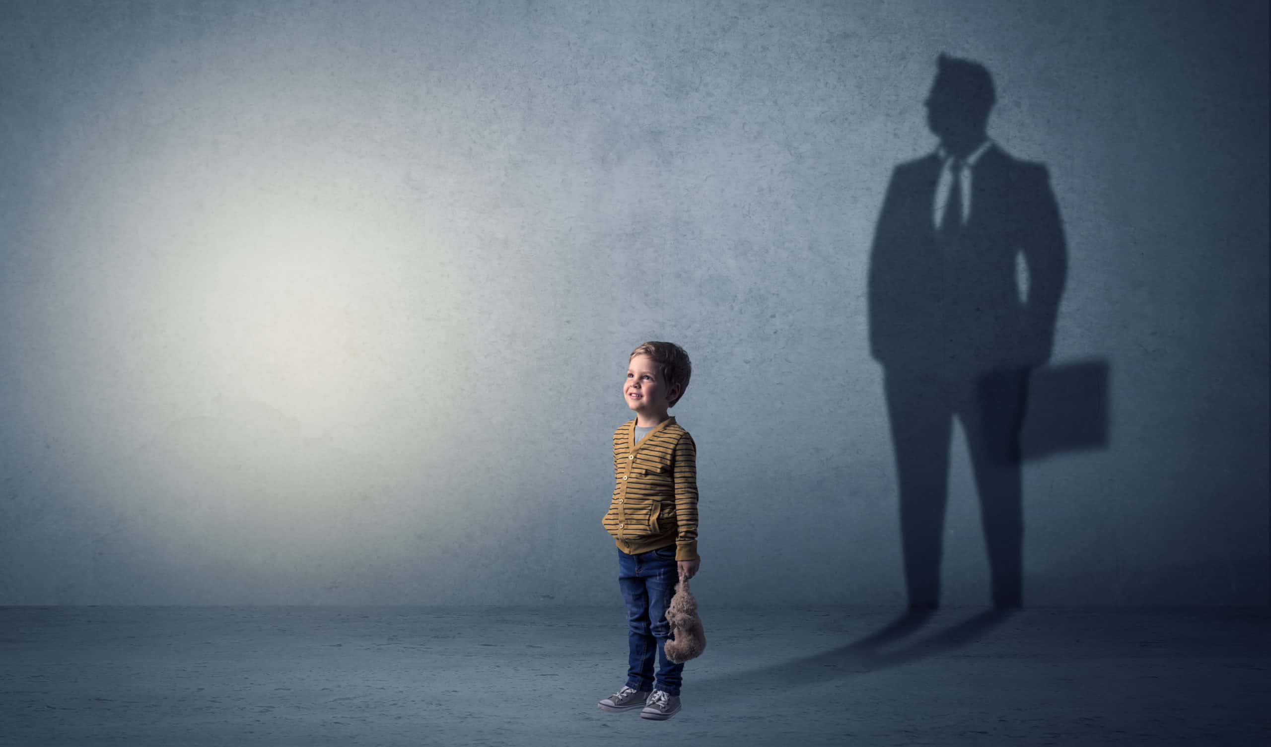 Kleiner Junge mit Teddybär in der Hand vor grauem Hintergrund. Sein Schatten zeigt einen erwachsenen Mann mit Anzug und Krawatte.