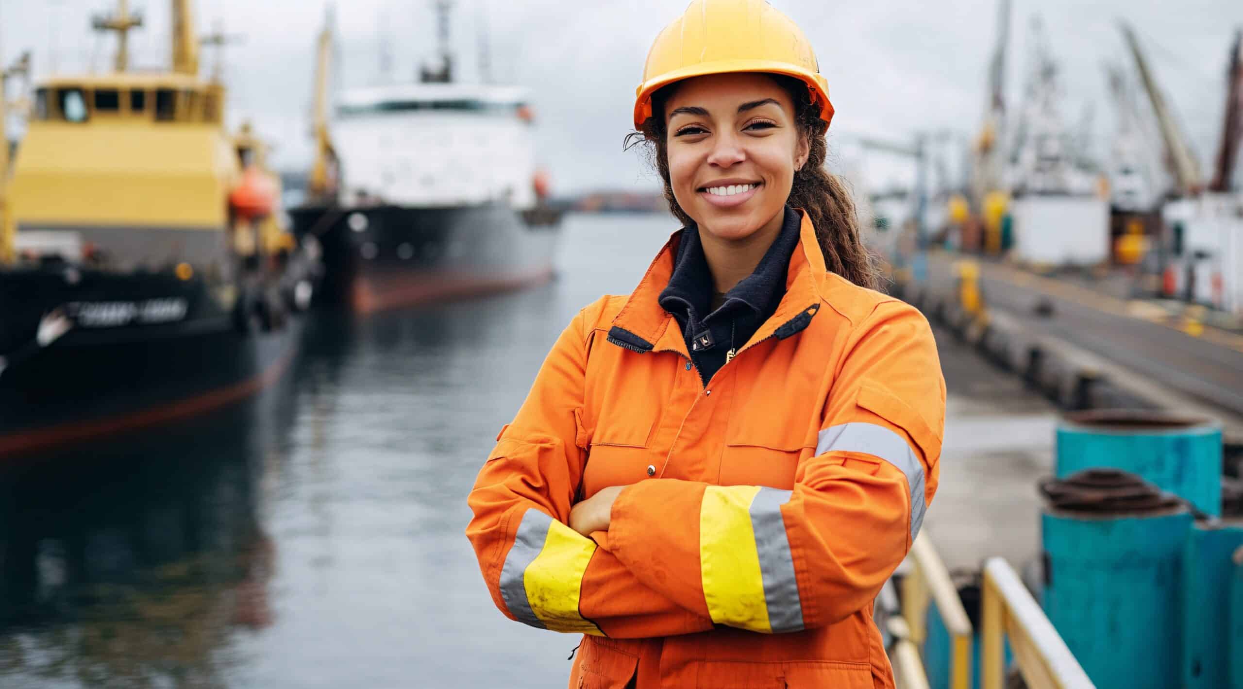 Female Engineer Overlooking Harbor Operations Frau mit Schutzhelm und orangener Sicherheitsjacke steht vor einem Hafen mit großen Schiffen