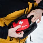 Person mit gelb-schwarzer Jacke holt ein kleines rotes Erste-Hilfe-Täschchen aus einer schwarzen Hüfttasche heraus, zu sehen sind nur der Oberkörper, die Hände und die beiden Taschen.