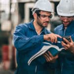 Zwei Männer im Blaumann mit weißem Schutzhelm und Schutzbrille stehen in einem industriellen Umfeld und schauen prüfend in ein großes Handbuch.