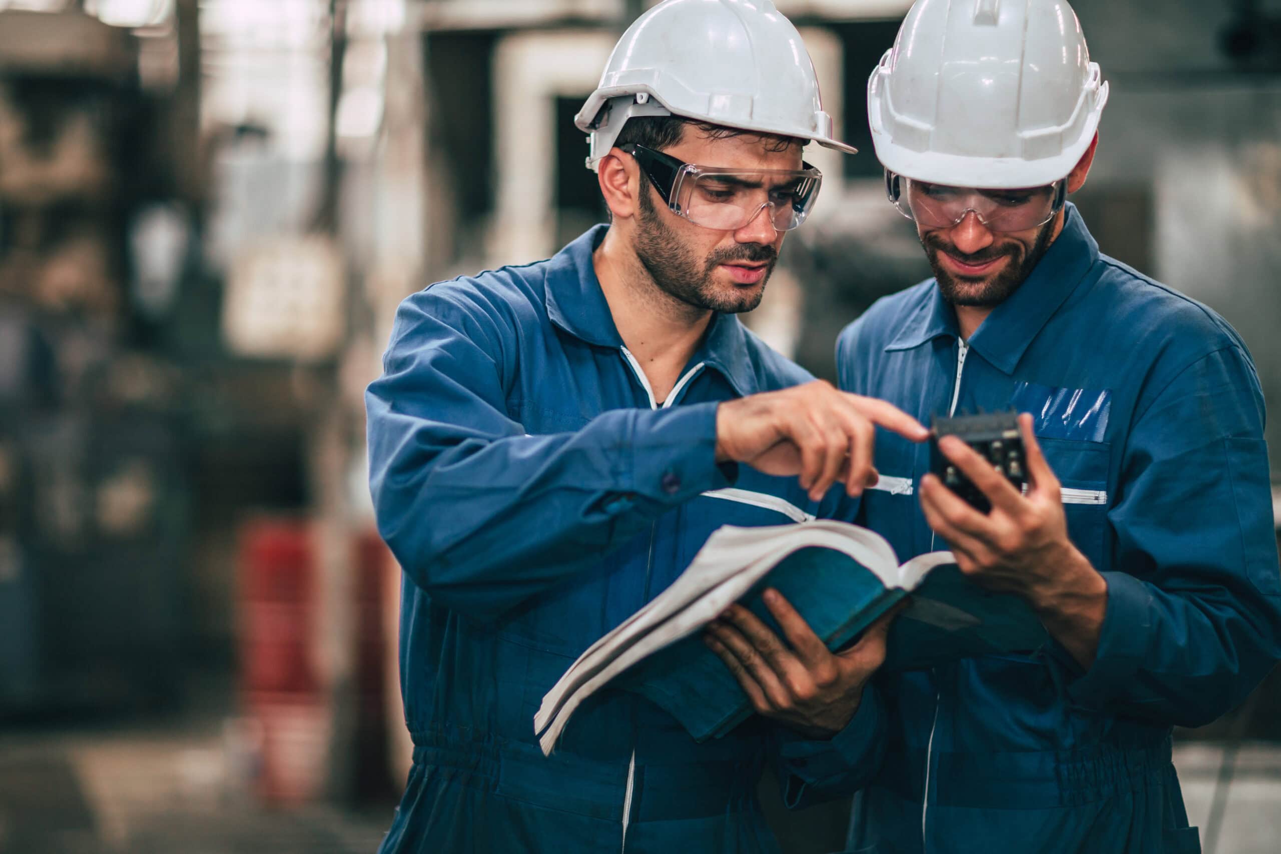Zwei Männer im Blaumann mit weißem Schutzhelm und Schutzbrille stehen in einem industriellen Umfeld und schauen prüfend in ein großes Handbuch.