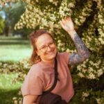 Anna, Mitarbeiterin von midok, steht vor einem blühenden grpßen Busch und lächelt in die Kamera.