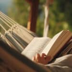 Blick auf ein Buch, dass eine Person in einer Hängematte auf dem Schoß liegen hat, im Hintergrund sind Bäume zu sehen.