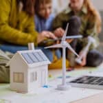 Im Vordergrund sieht man Modelle von Solaranlagen auf einem Hausdach und ein Windrad in Form von Holzspielzeug, unscharf im Hintergrund sitzen 2 erwachsene Personen mit 2 Kindern und zeigen etwas auf einem Tablet