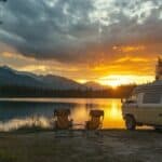 Ein Camper steht an einem See im Wald. Daneben stehen zwei Klappstühle und zusehen ist der Sonnenuntergang mit Wolken am Himmel.