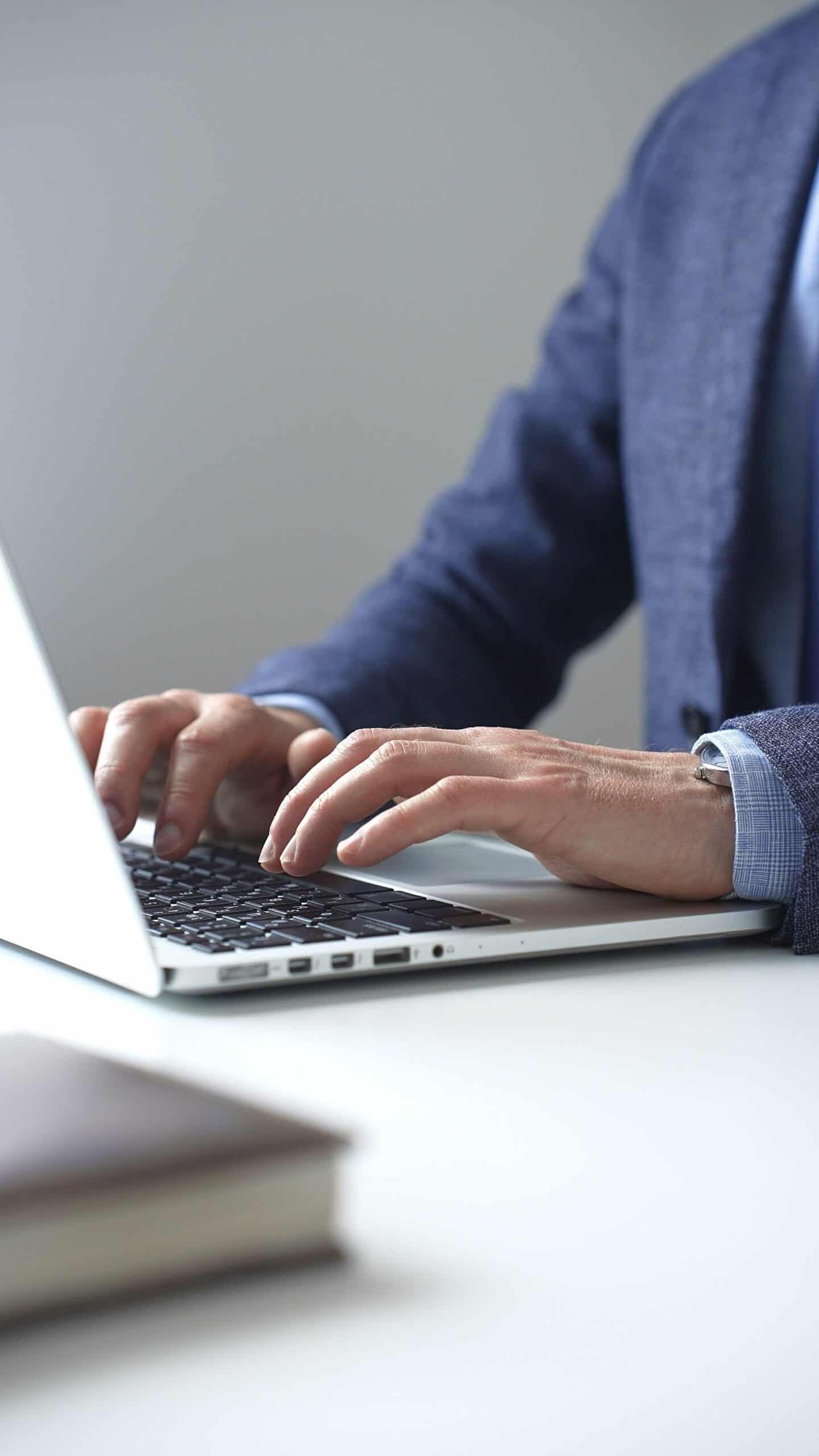 Vertikale Nahaufnahme der Hände eines Geschäftsmannes über einer Laptop-Tastatur. Er trägt einen dunkelblauen Anzug und eine Uhr. Unscharf im Vordergrund liegt ein Buch.