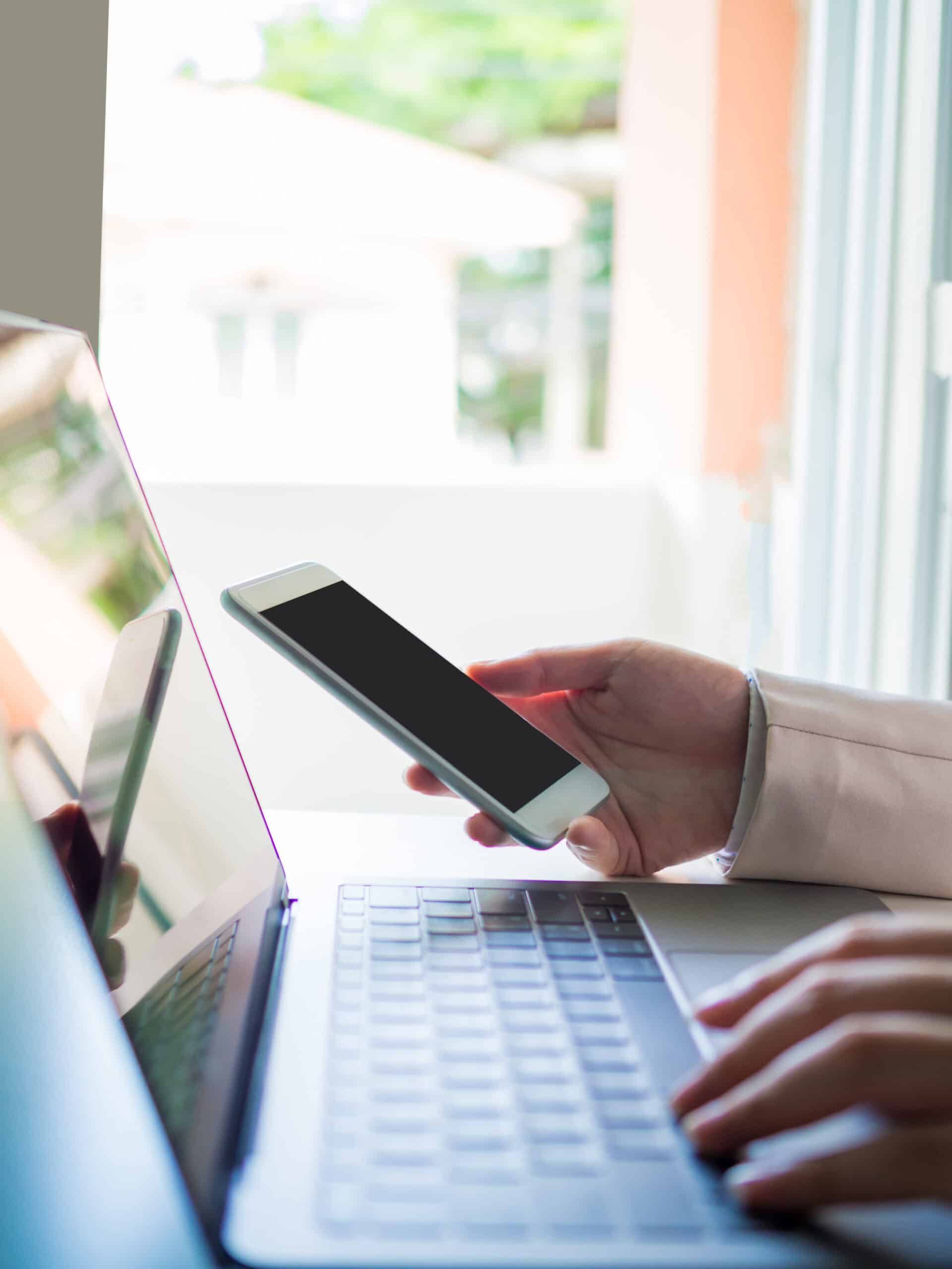 Nahaufnahme eines Smartphones in der rechten Hand einer Geschäftsfrau in einem pastellfarbenen Anzug, die in ihrem Homeoffice mit ihrem Laptop arbeitet, mit weichgezeichneter linker Hand und Tastatur im Vordergrund.