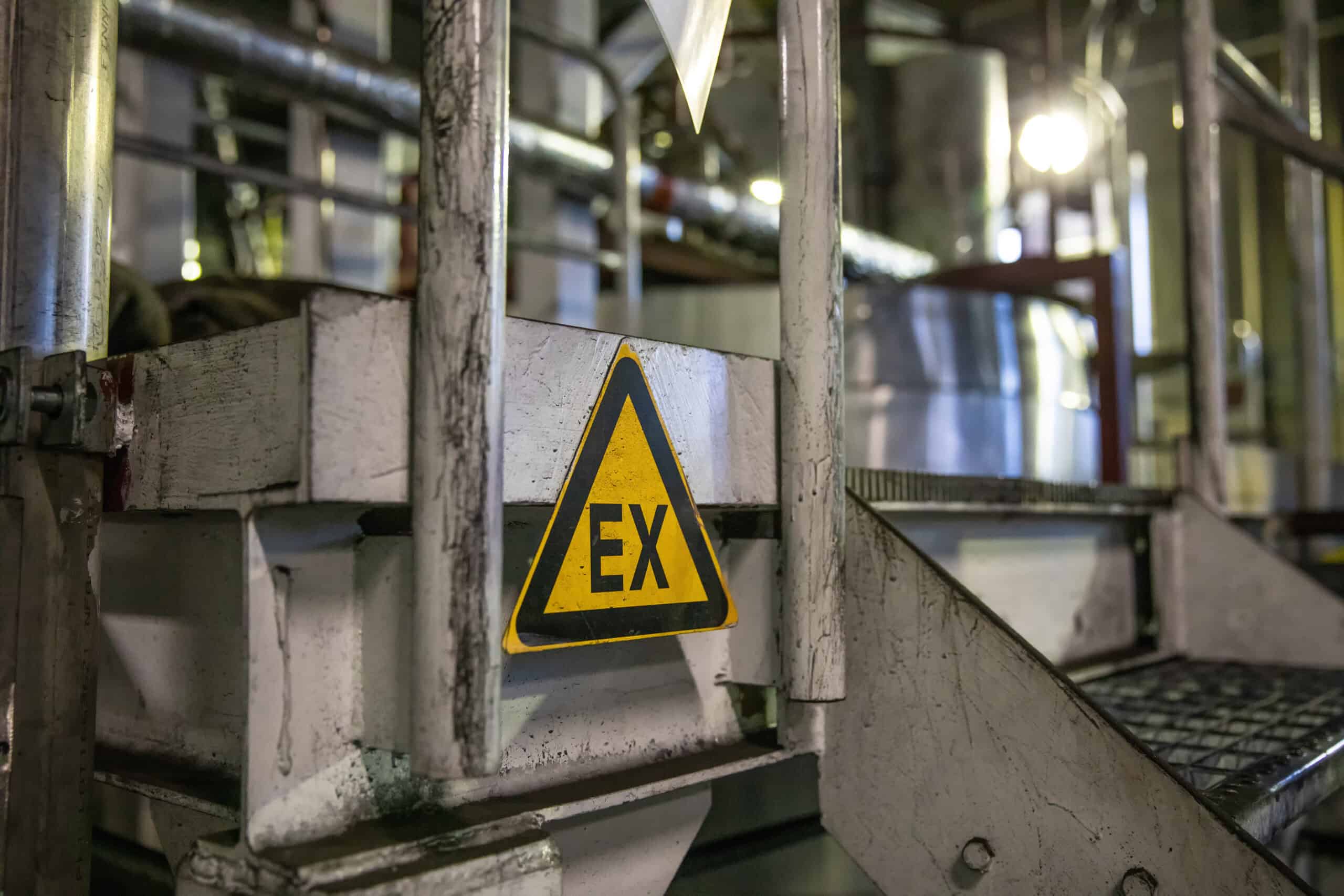 Nahaufnahme eines Warnschilds für explosionsfähiger Atmosphäre, welches am Geländer einer alten Stahltreppe hängt.