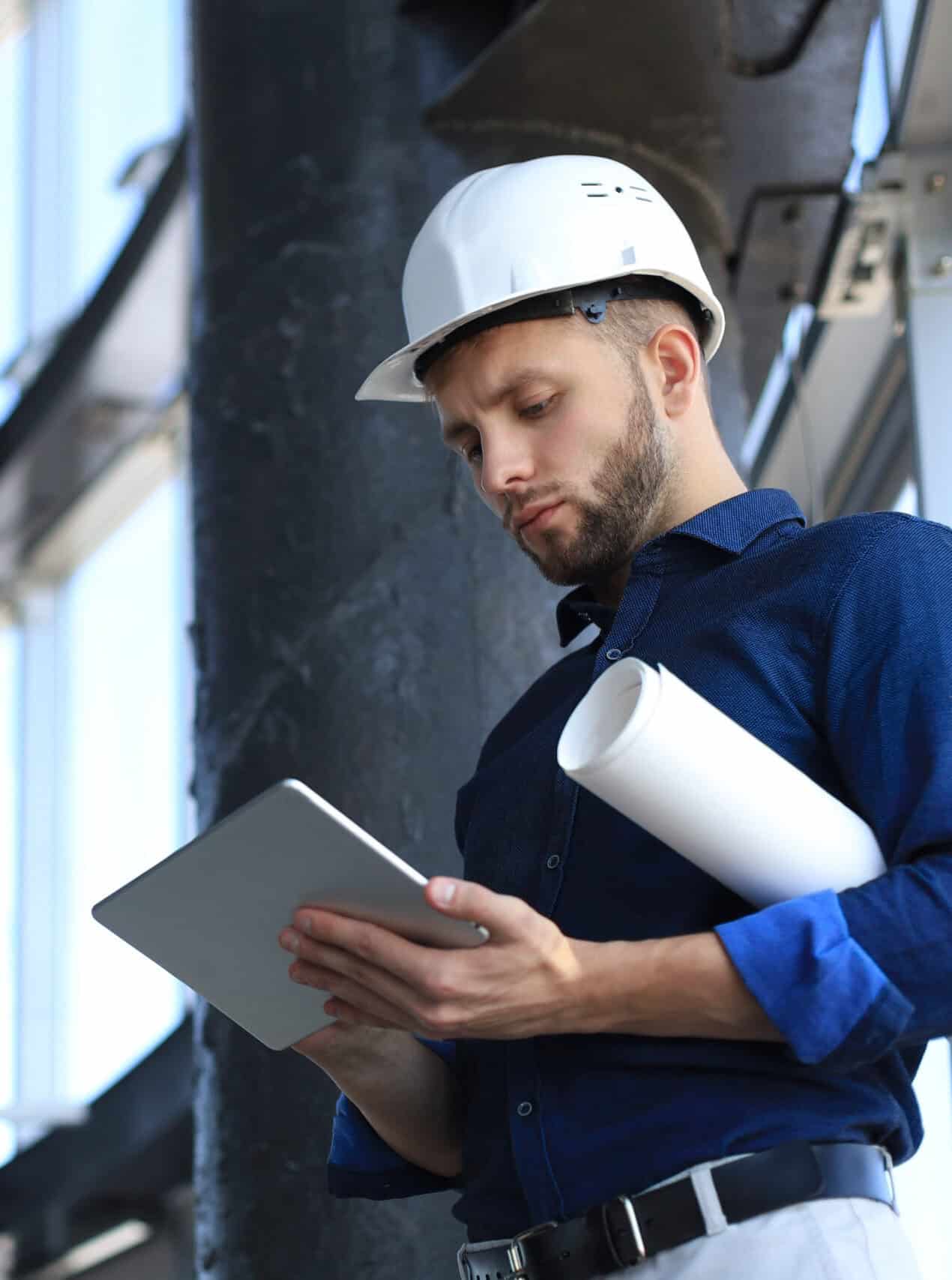 Ein Mann im blauen Hemd und mit weißem Helm steht vor einer Fensterfront in einem leeren Gebäude. In der Hand hat er ein Tablet, worauf er schaut.