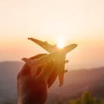 Eine Hand hält ein Spielzeug-Flugzeug in den Himmel. Im Hintergrund sind Berge mit Sonnenuntergang zu sehen.